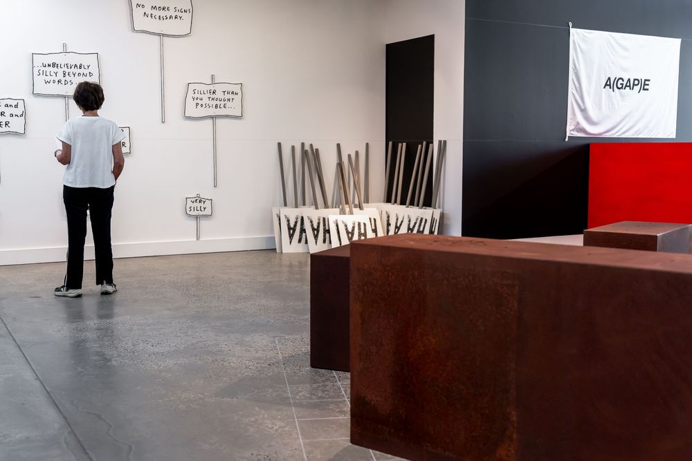Person viewing protest sign artwork installation in contemporary art gallery with rusty steel sculptures.