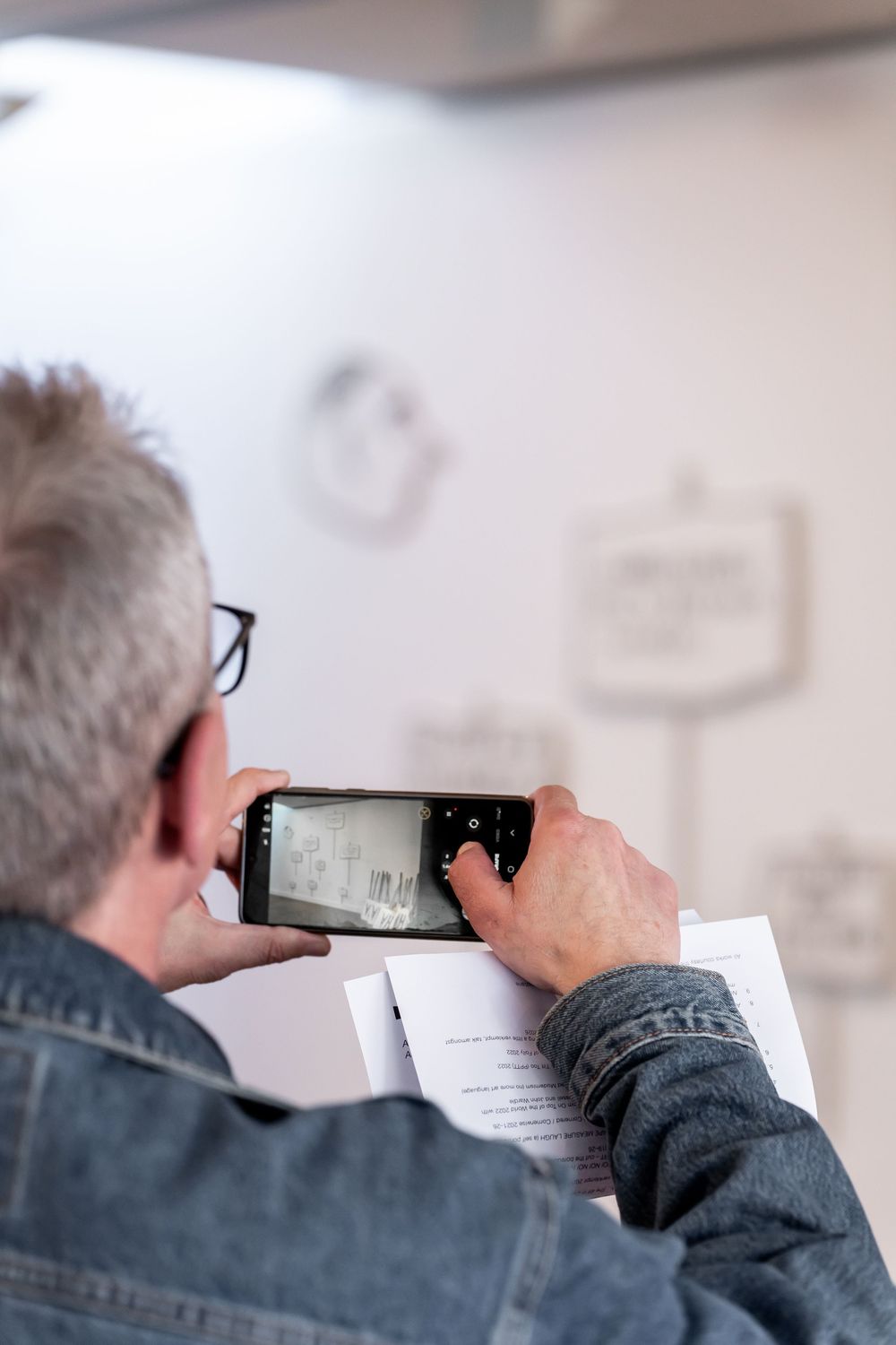 Man in denim jacket photographing a document with his smartphone in a modern white office space.