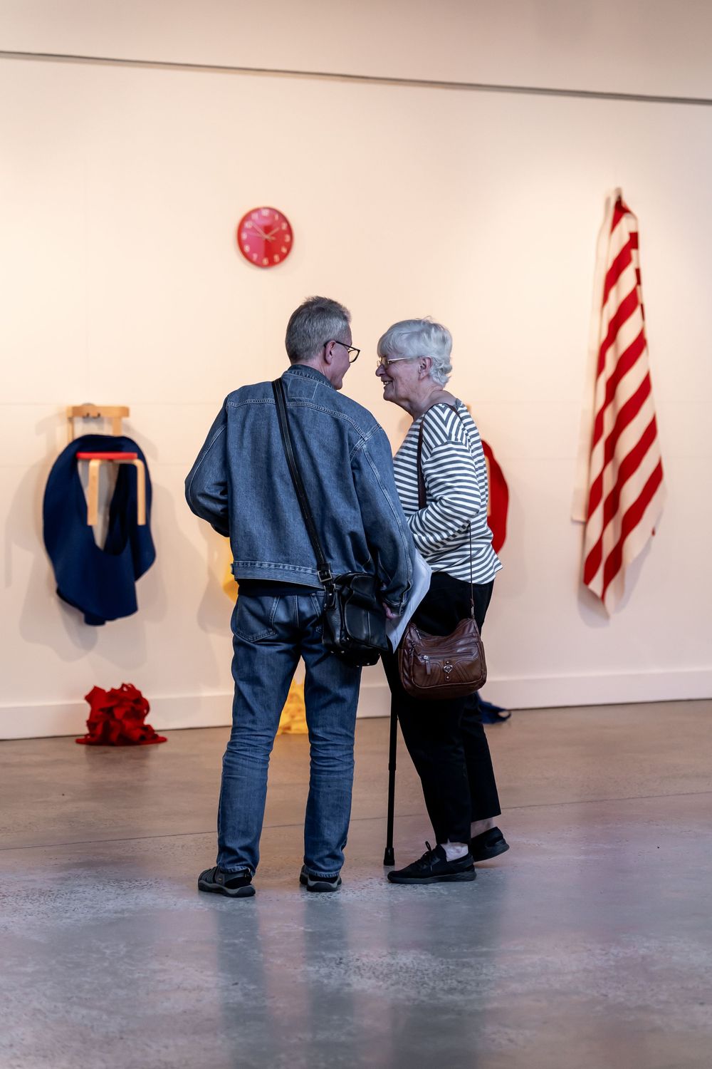 Two visitors converse in a modern art gallery surrounded by colorful textile installations on white walls.