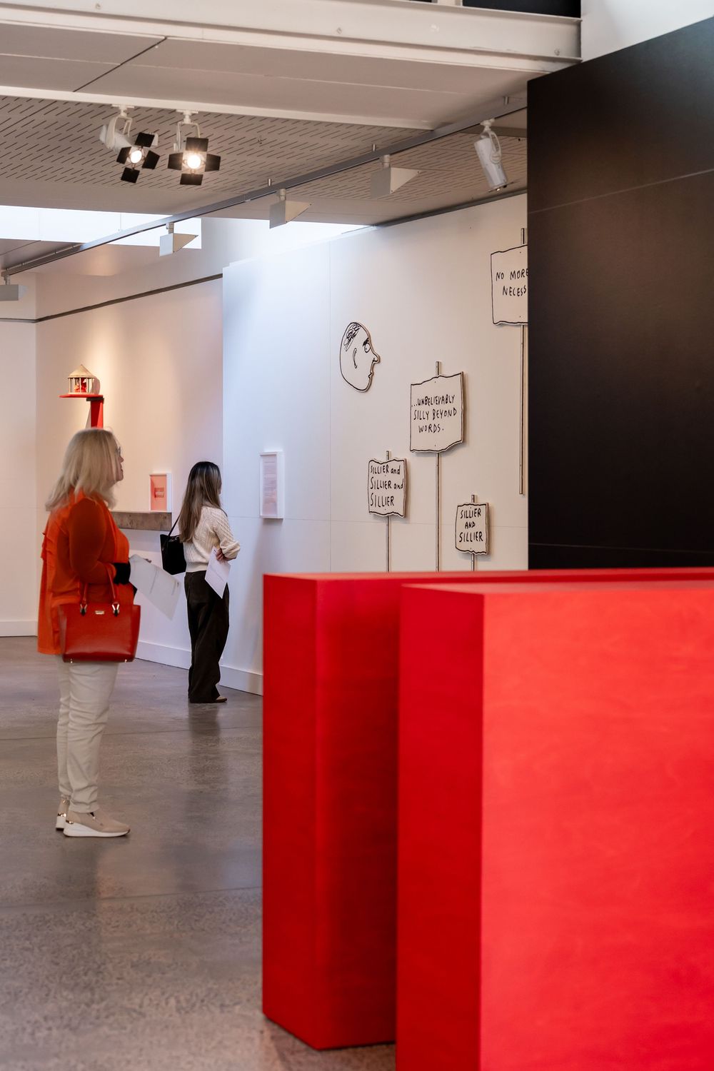 Two women viewing artwork in a modern gallery with red display fixtures and illustrated signs on white walls.