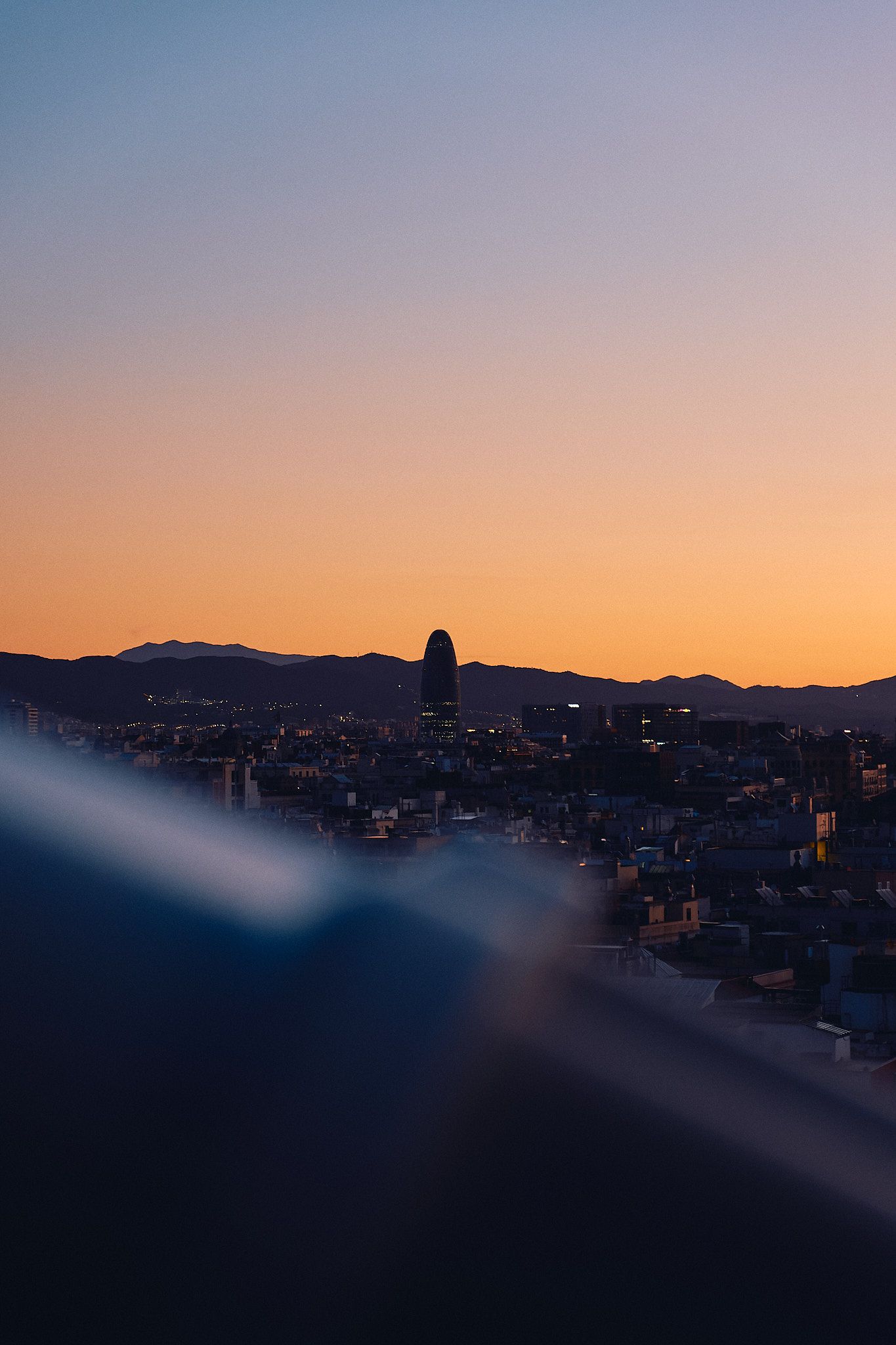 Aerial cityscape at dusk with a prominent tower silhouetted against a warm orange sunset sky and distant mountains.