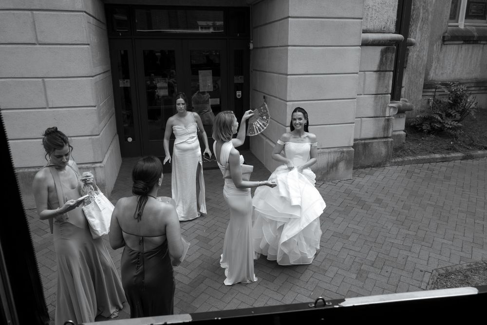 Black and white photo of bridesmaids in elegant gowns gathered outside a building, preparing for a wedding ceremony.