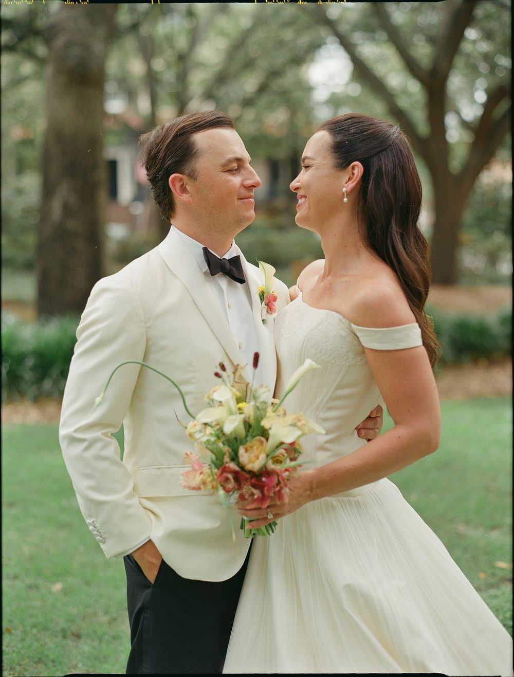 A bride and groom smile at each other outdoors, she holds a floral bouquet, he wears a cream tuxedo jacket with bow tie.