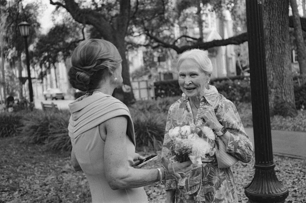 Two women conversing outdoors in a park, one holding flowers, captured in black and white photography.