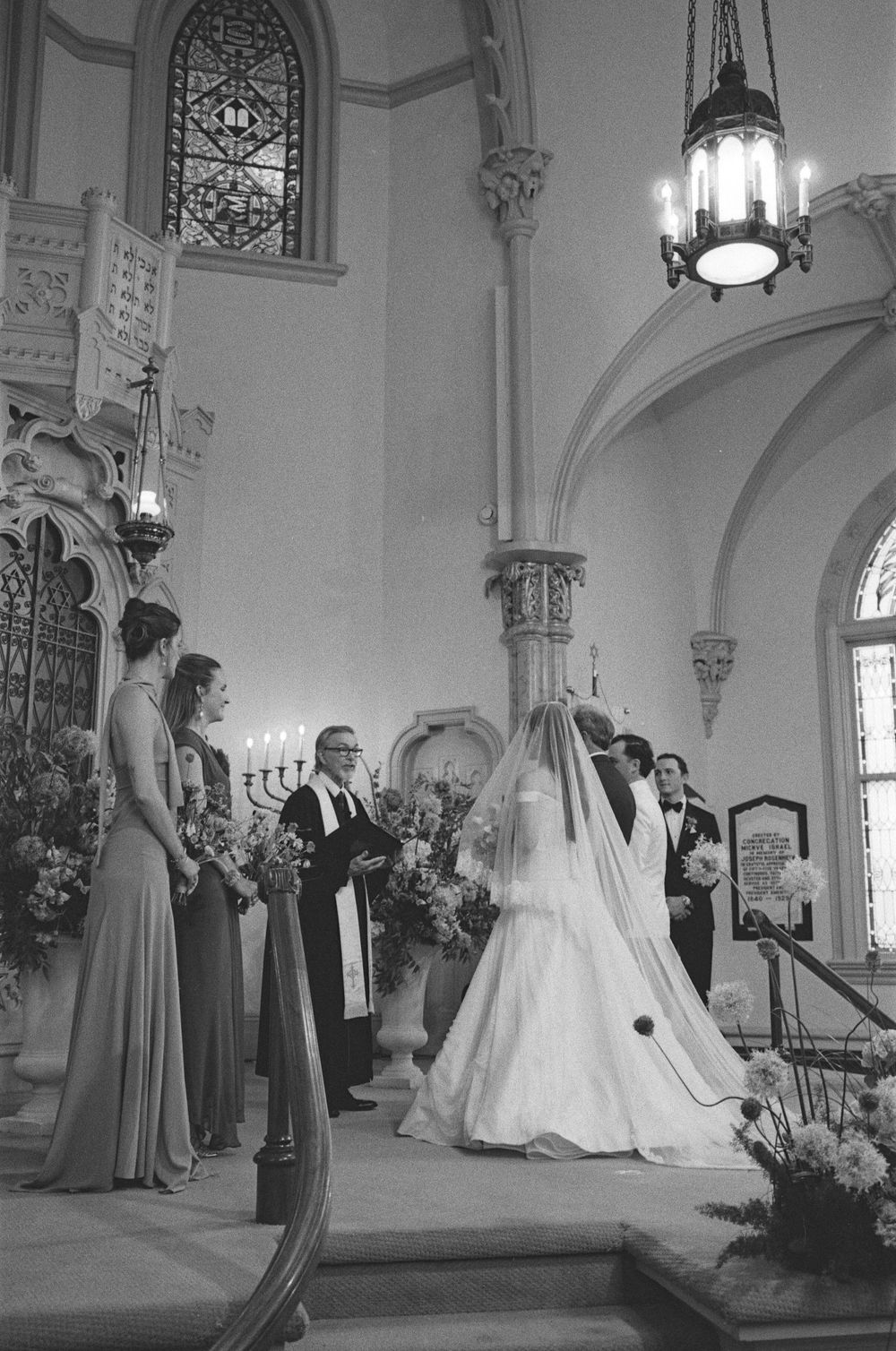 Black and white photo of a bride and groom exchanging vows at a church altar with bridesmaids and officiant.