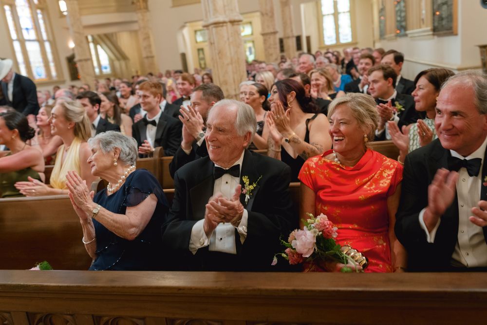 Joyful wedding guests clapping and smiling in a grand church, dressed in formal attire including a red dress.