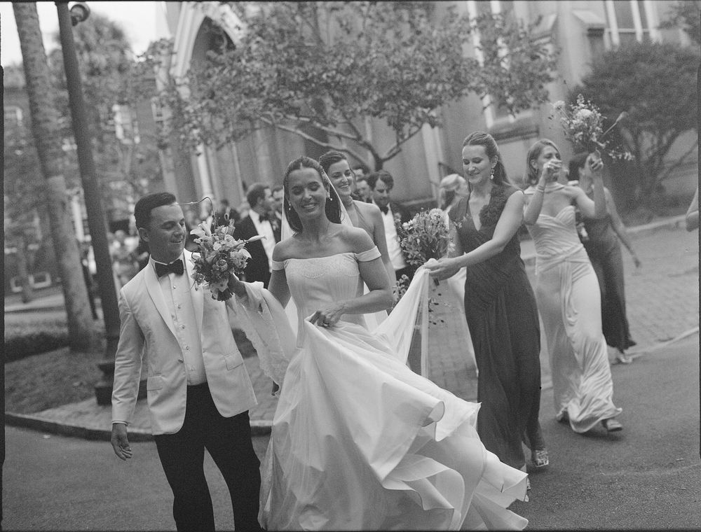 Black and white photo of a bride in a white gown walking with groom and bridesmaids outside a church.