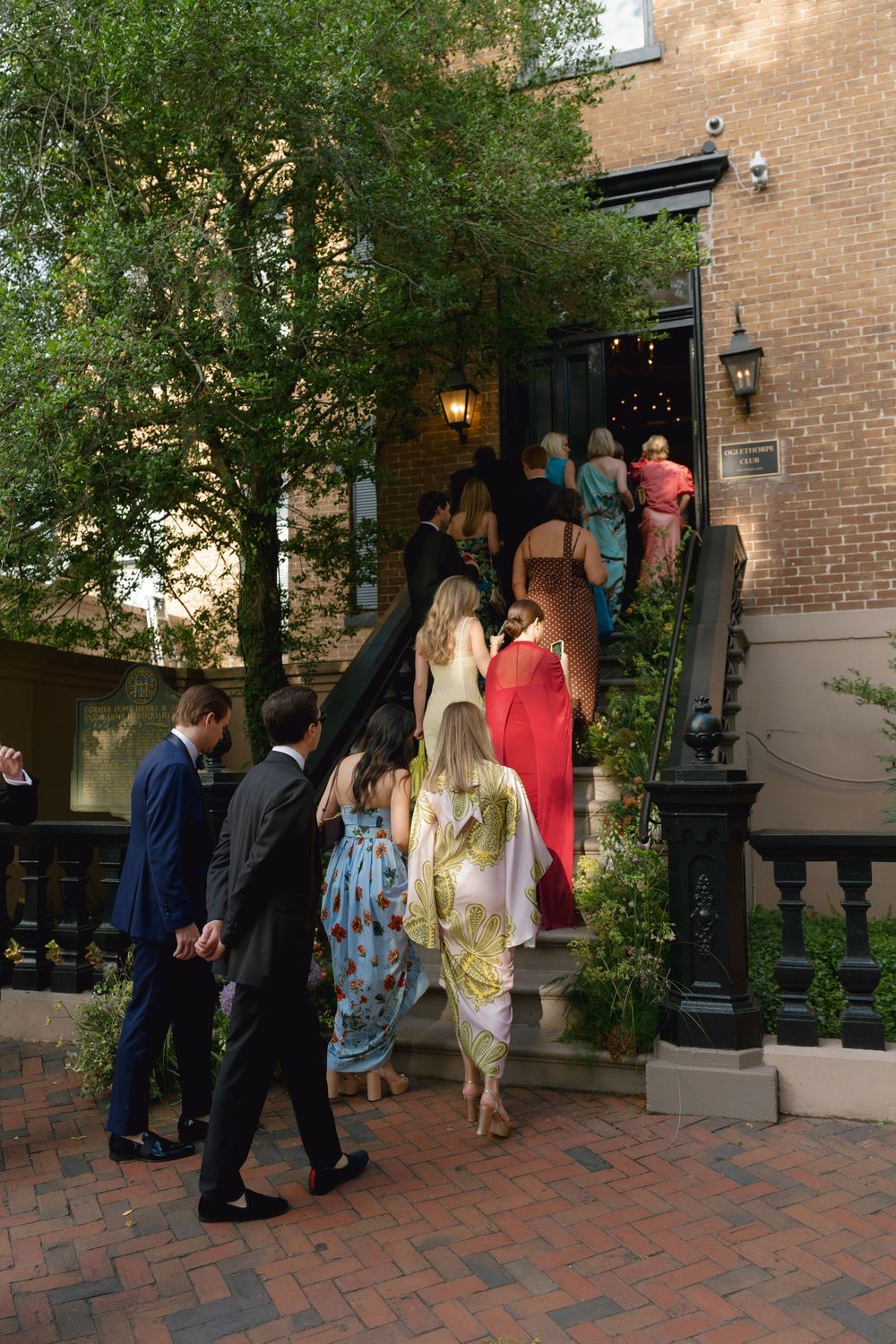 Elegantly dressed wedding guests ascending brick steps into a historic venue surrounded by lush green trees.