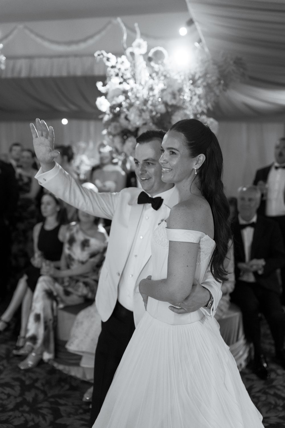 Bride and groom share first dance in black and white photo, groom waving in white tuxedo jacket.