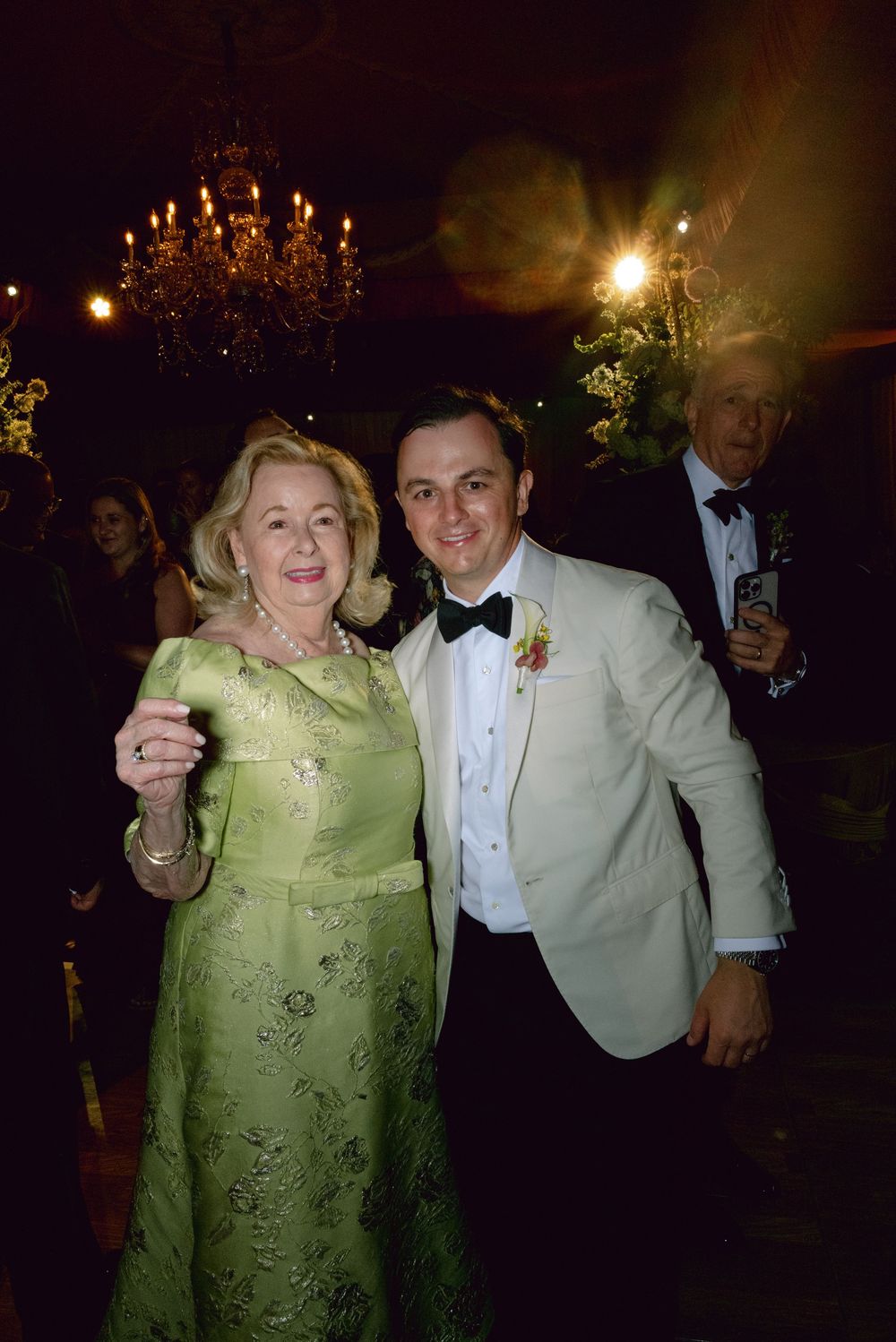 Elderly woman in green gown and young man in white tuxedo posing together at elegant formal event with chandelier.
