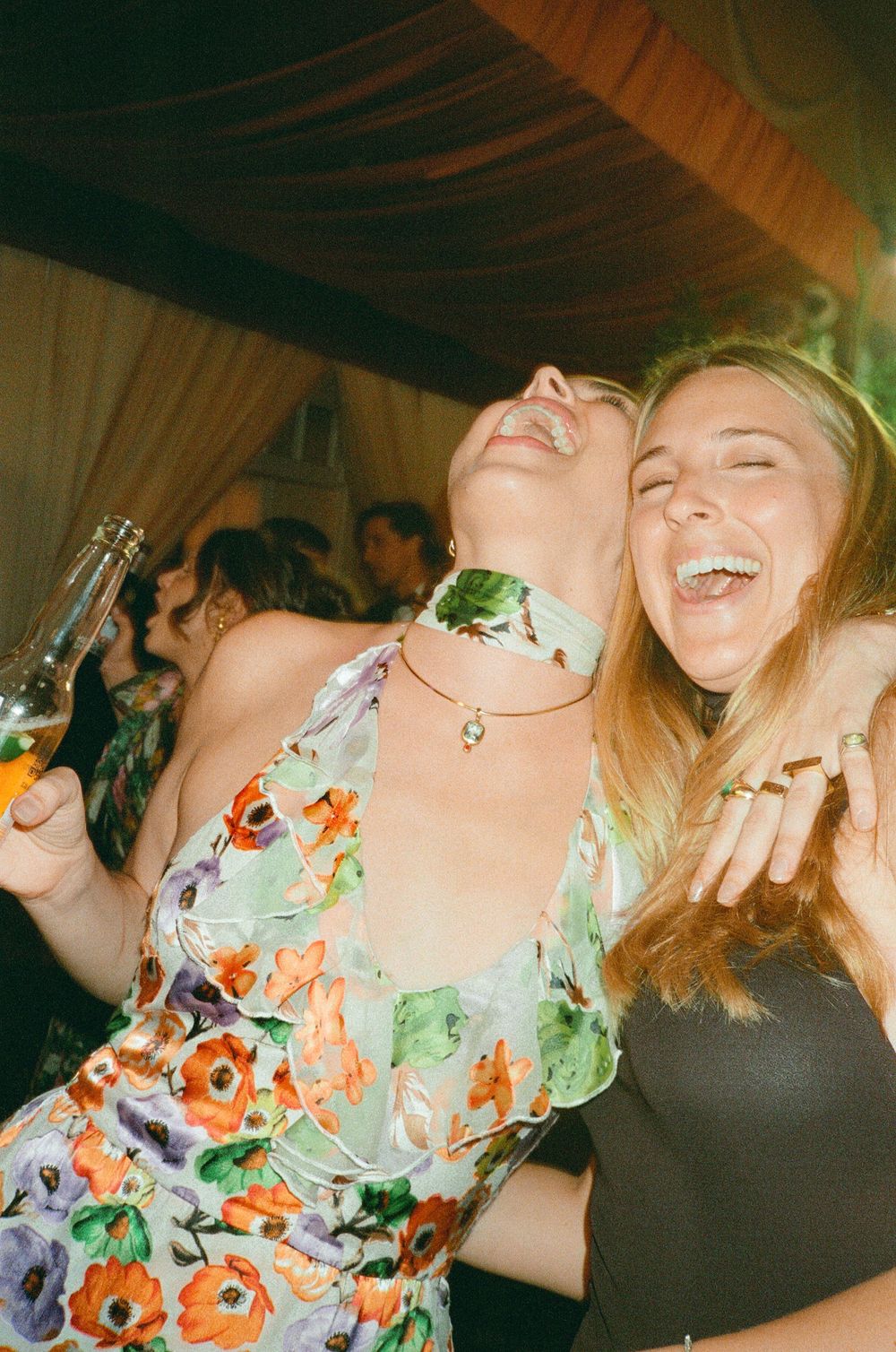 Two women laughing and dancing at a party, one wearing a floral dress holding a beer bottle.