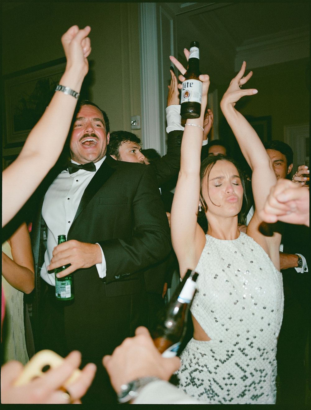 Guests in formal attire celebrating at a party, raising beer bottles with arms up, dancing joyfully indoors.