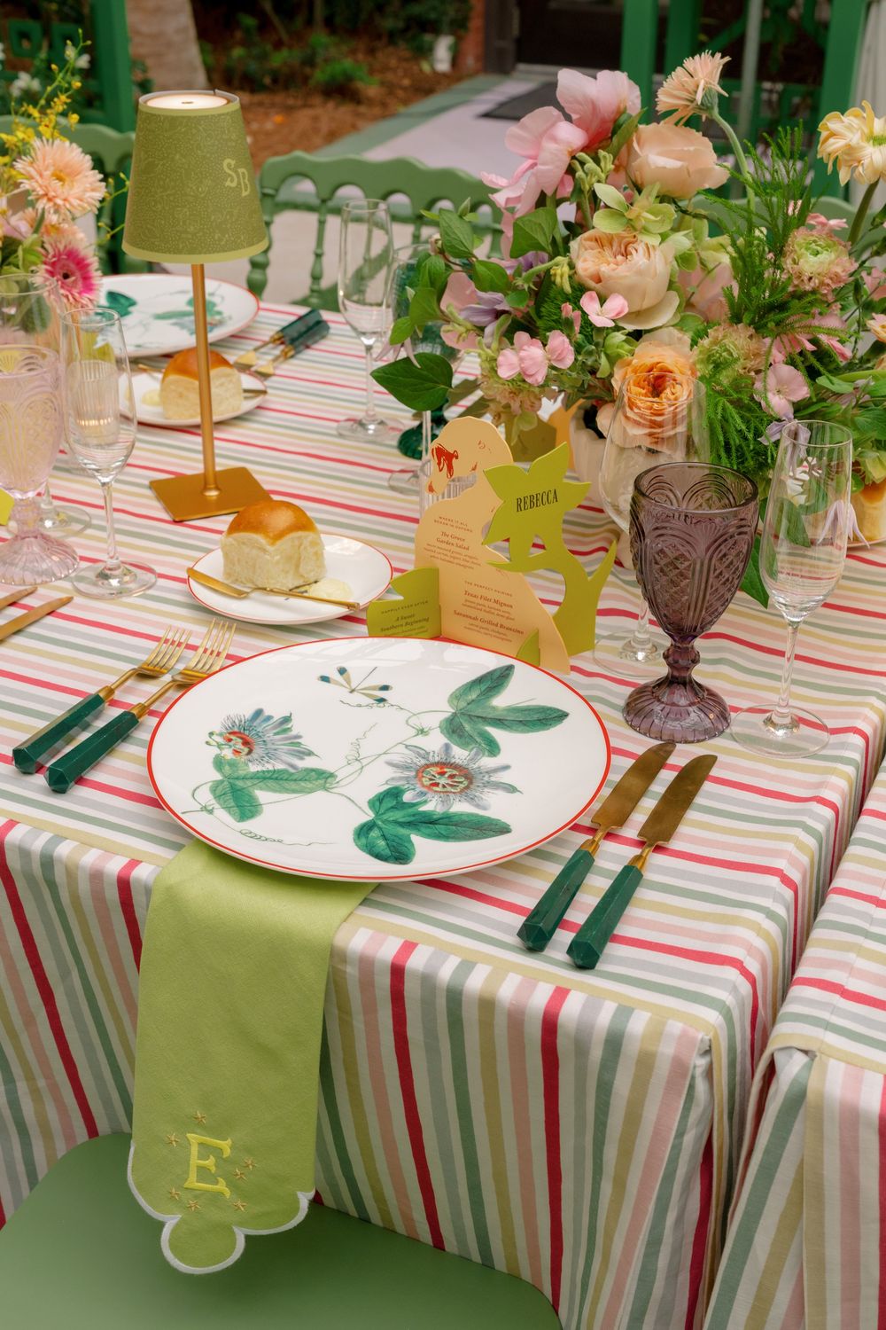 Elegant garden wedding tablescape with striped linens, botanical plates, gold cutlery, pink floral centerpiece, and green lamp.