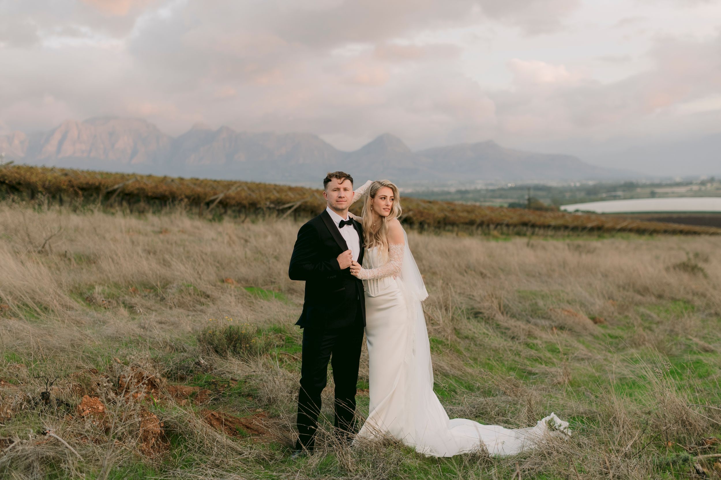 Bride and groom pose in a scenic field with mountain backdrop at sunset, bride in white gown, groom in black tuxedo.