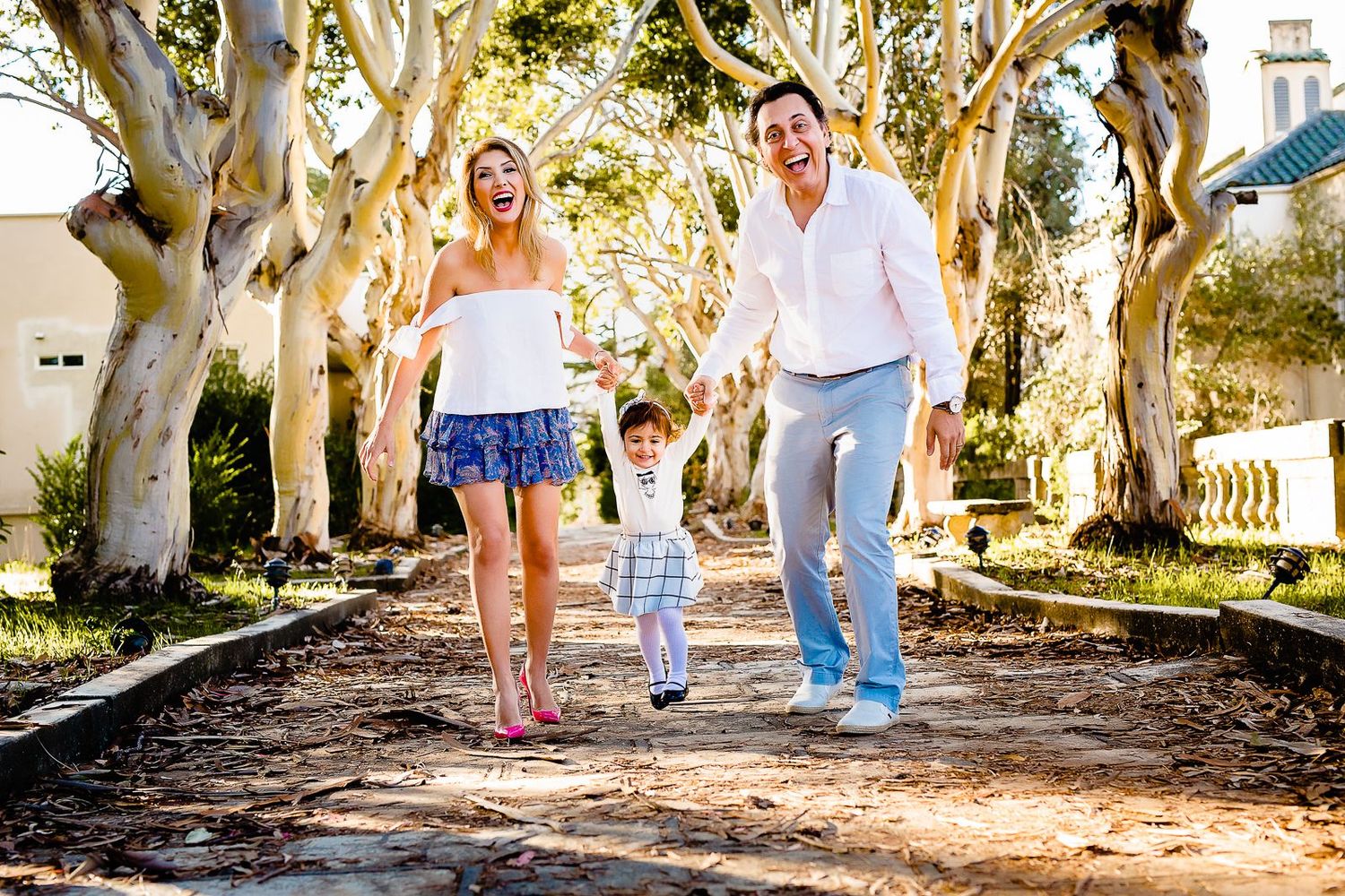 Happy family with young child swinging between parents on a sunny tree-lined path outdoors.