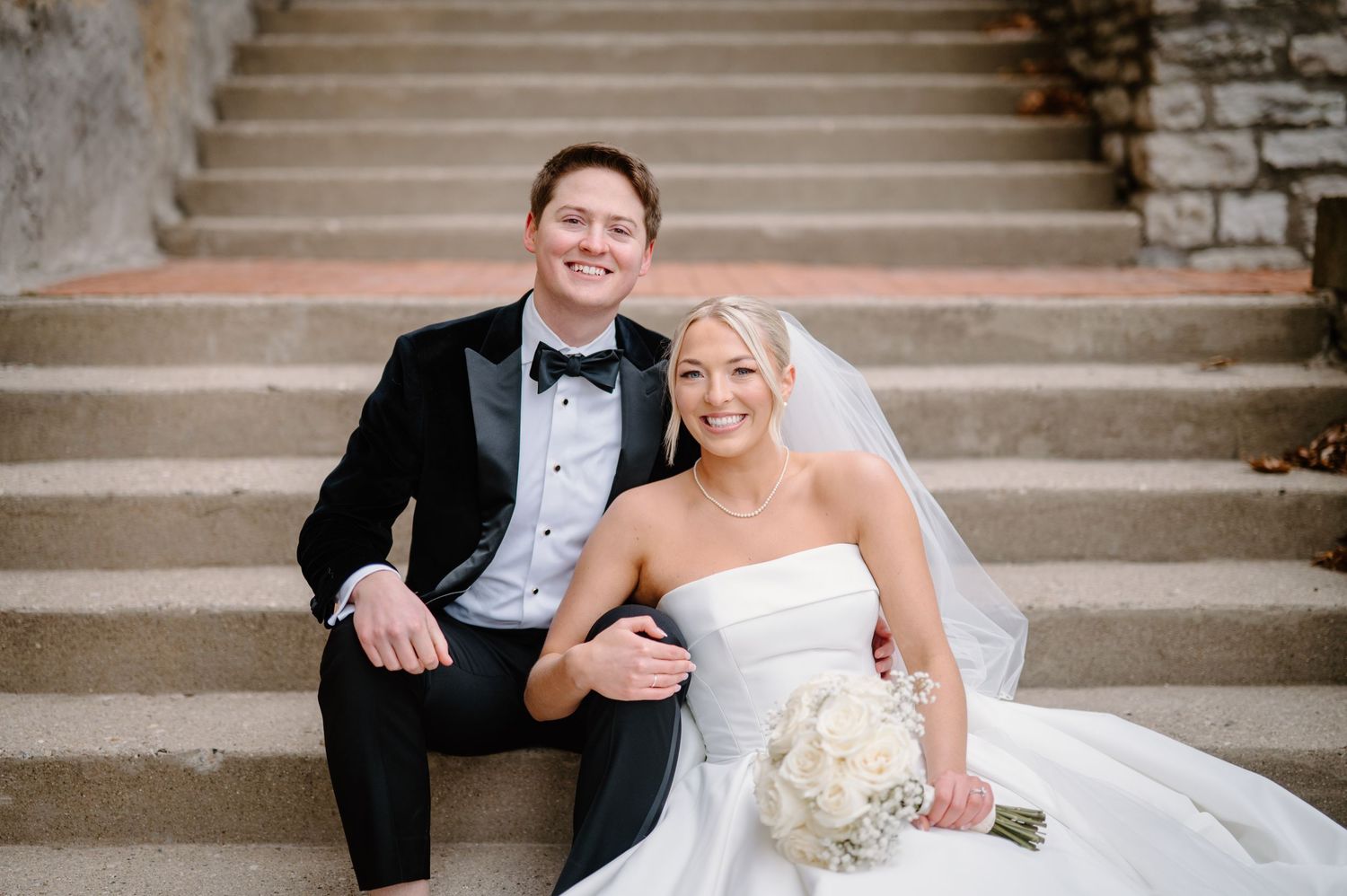 Bride and groom embrace near a tree by a pond, wedding veil flowing in the wind on a sunny day.
