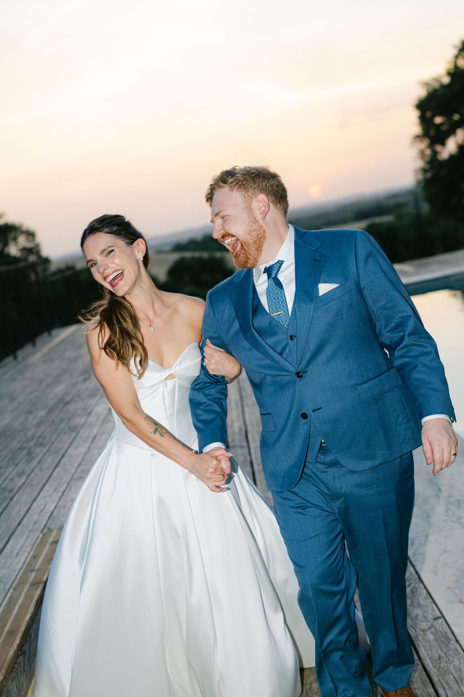 Bride and groom dancing on wooden deck at sunset near pool, bride in white gown holding champagne, groom in blue suit.