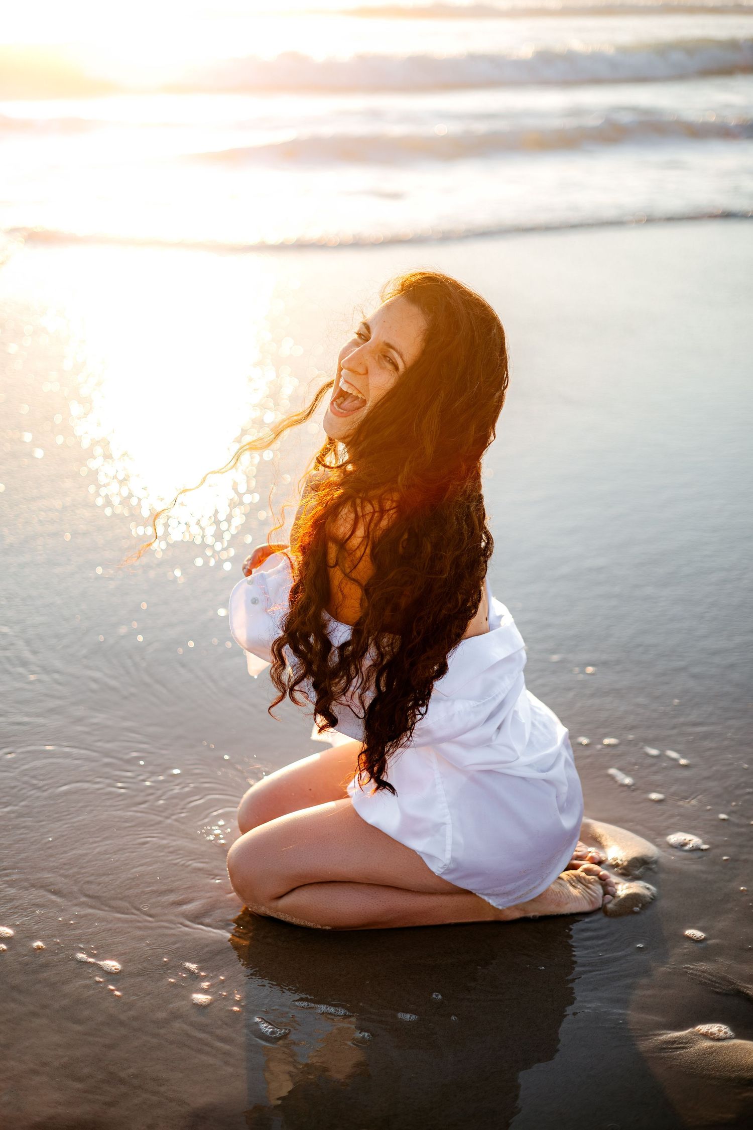 Jeune femme aux longs cheveux noirs et bouclés riant assise sur une plage mouillée au coucher du soleil.