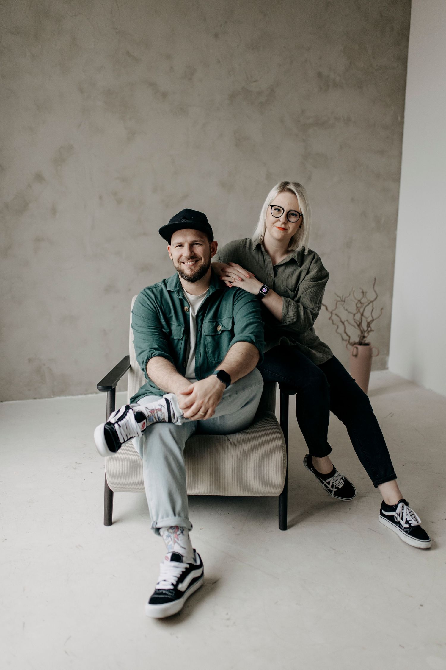 Stylish couple posing in modern studio, man in cap seated in chair with woman beside him.