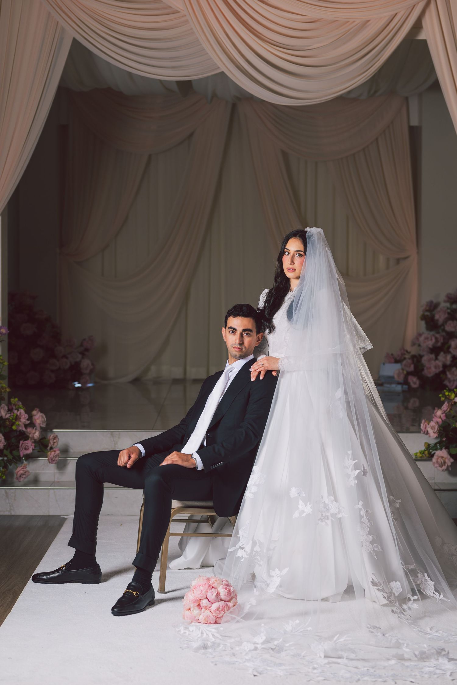 Elegant bride in white gown with veil stands beside groom in black suit seated in chair at decorated wedding venue.