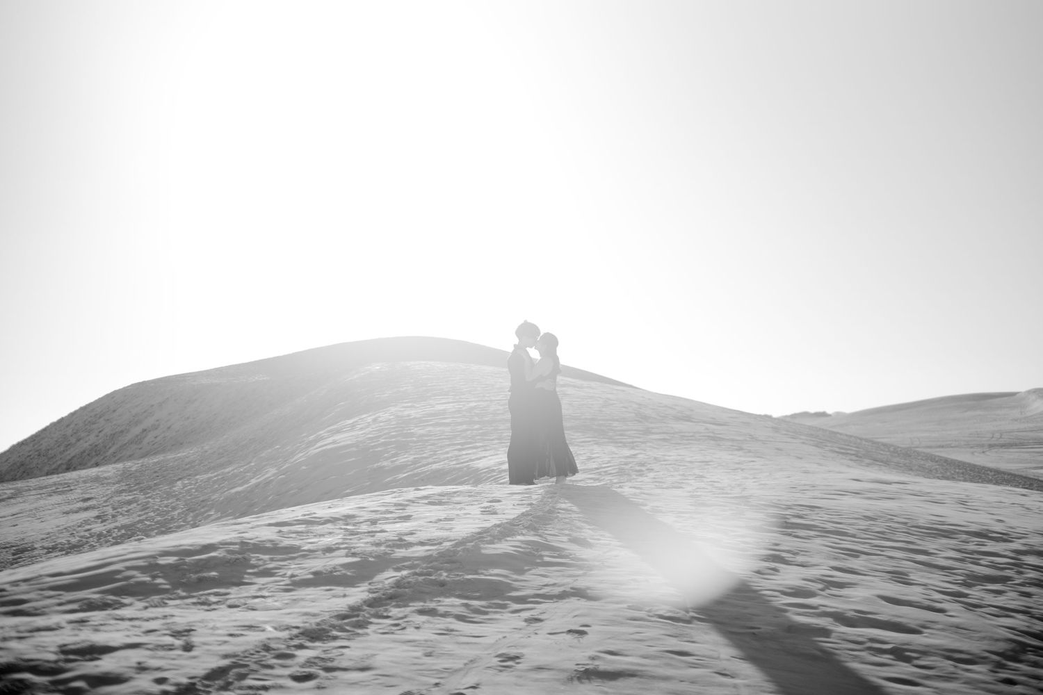 Black and white photo of a couple kissing on a snowy sand dune with bright sunlight behind them.