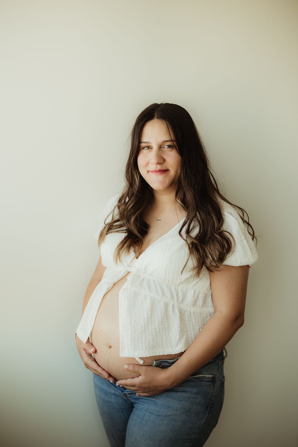 Pregnant woman in white crop top and jeans cradling baby bump, smiling softly against a light neutral background.