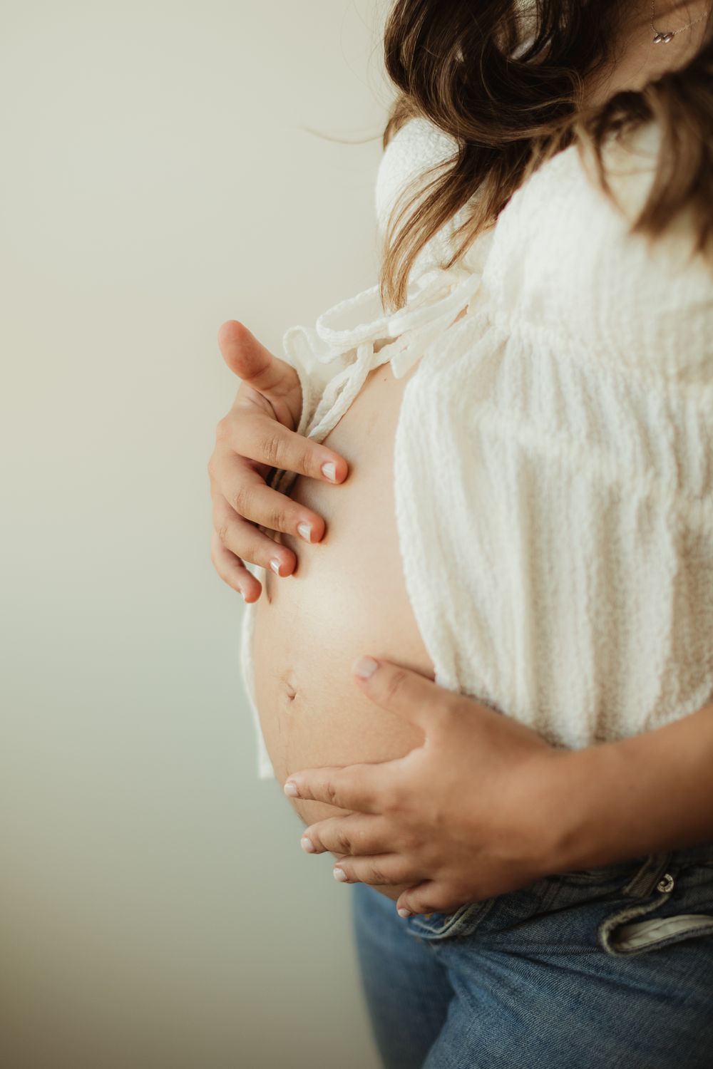 Pregnant woman in white off-shoulder top and jeans cradling her baby bump with both hands against a neutral background.
