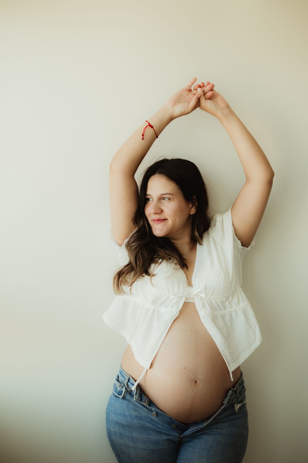 Smiling pregnant woman in white crop top and jeans, arms raised overhead, showing baby bump against light wall.
