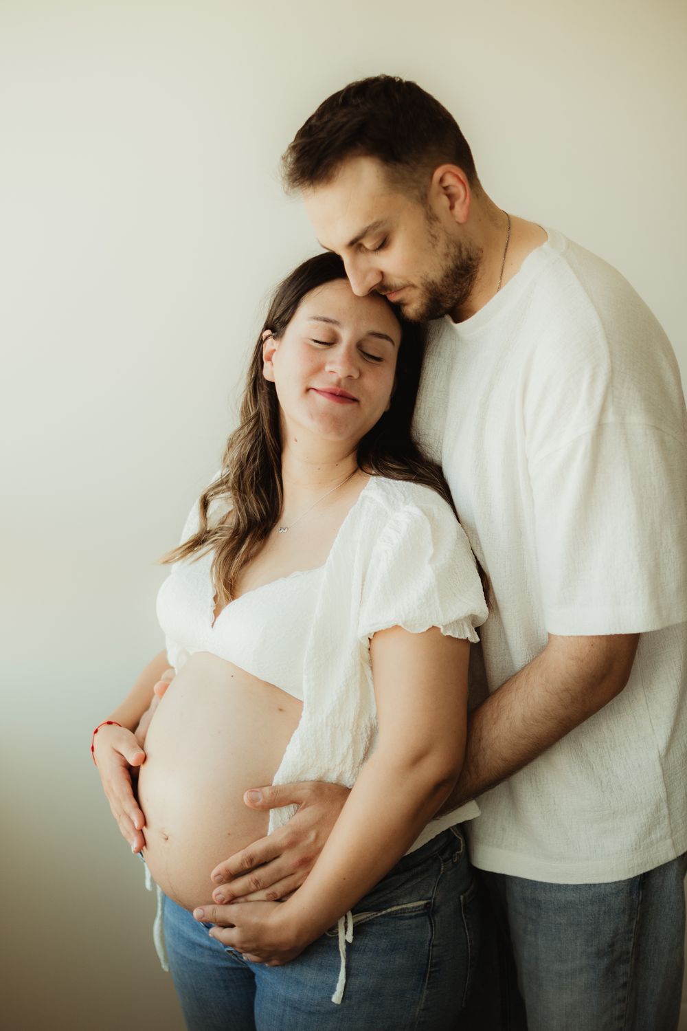 A couple in white tops tenderly embrace, the woman visibly pregnant, both smiling softly with eyes closed.