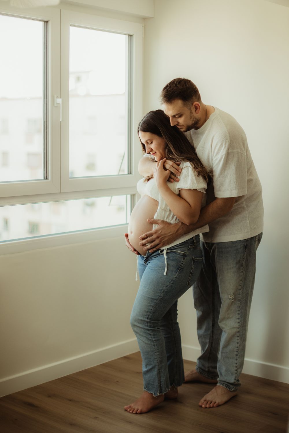 A couple shares a tender moment, man embracing pregnant woman from behind near a bright window in a modern room.