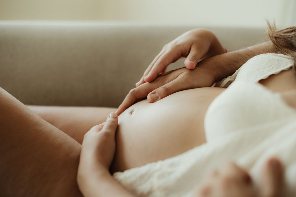 Pregnant woman in white lingerie gently cradling her baby bump while resting on a couch.