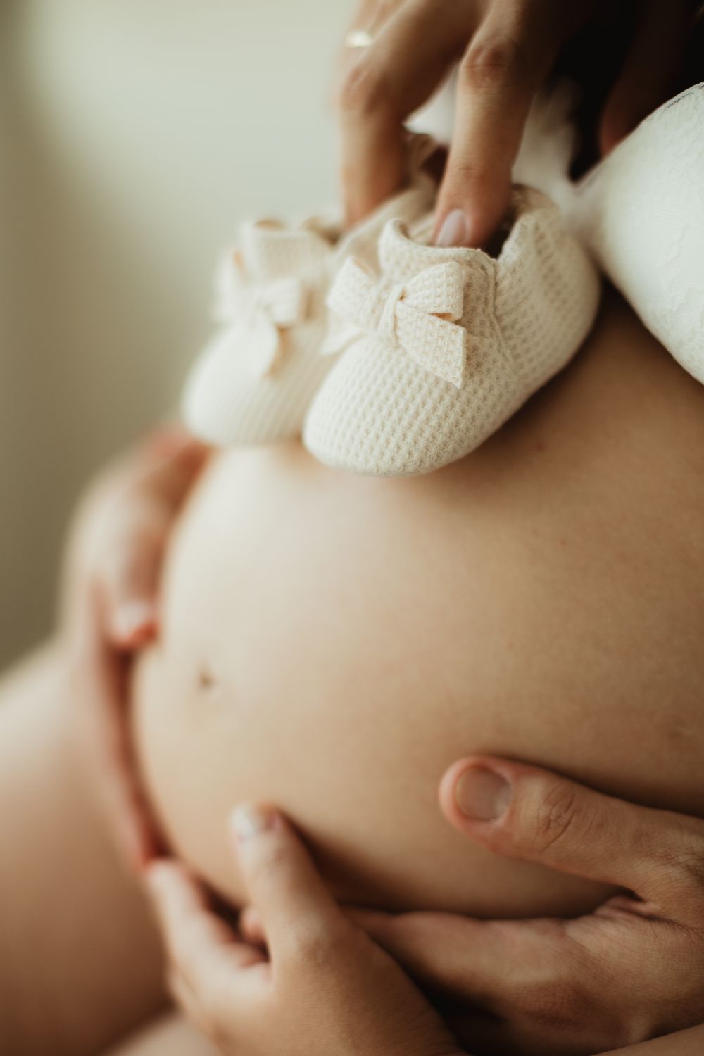 Pregnant woman cradling baby bump with tiny white knit booties with bow resting on top.
