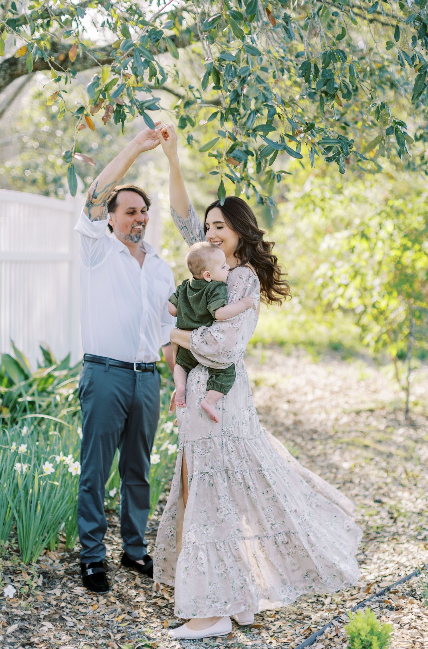 A joyful family of three poses under a tree, with a man, woman in a floral gown, and baby in a garden.