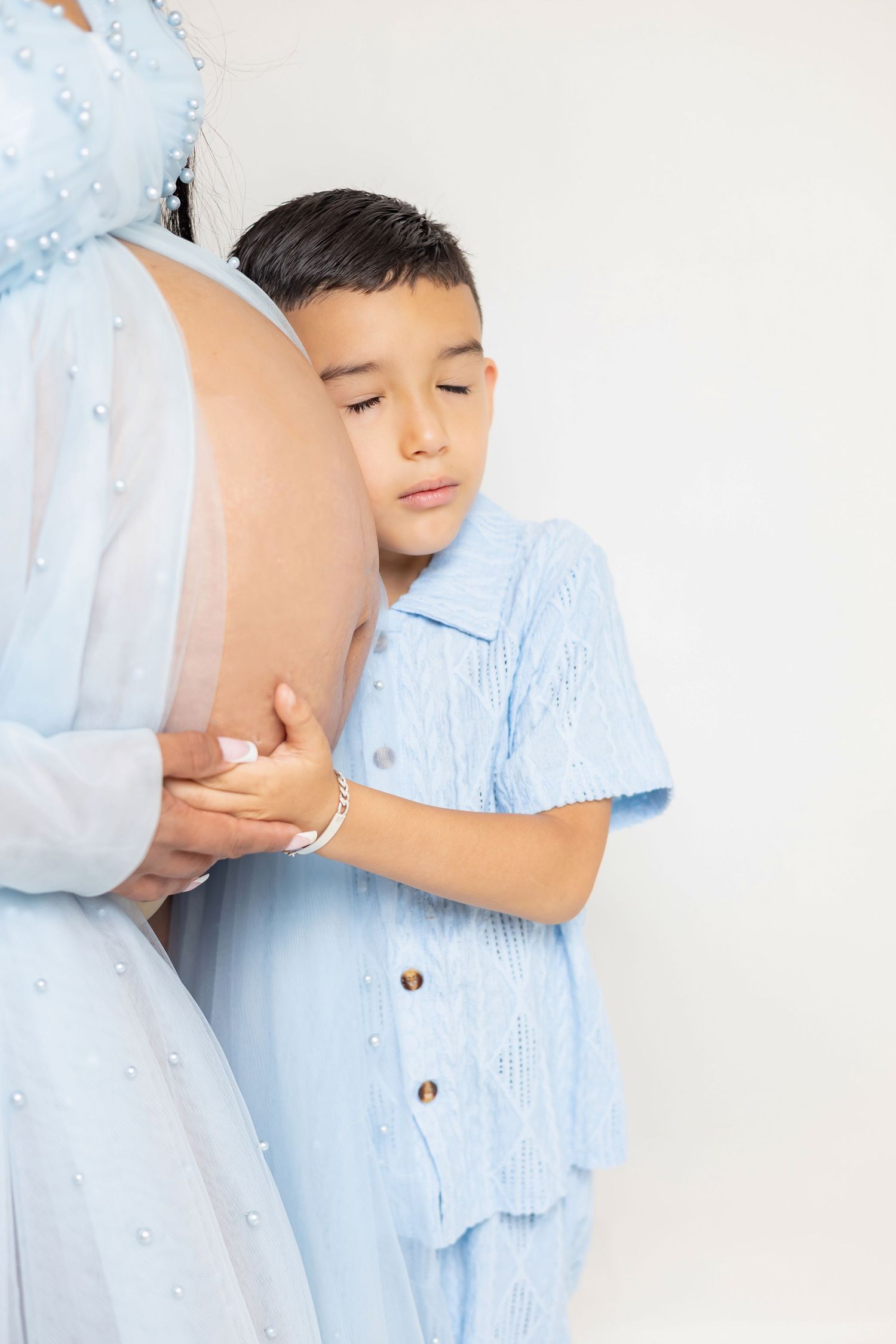 A young boy in blue shirt tenderly hugs his pregnant mother's belly, both dressed in matching light blue outfits.