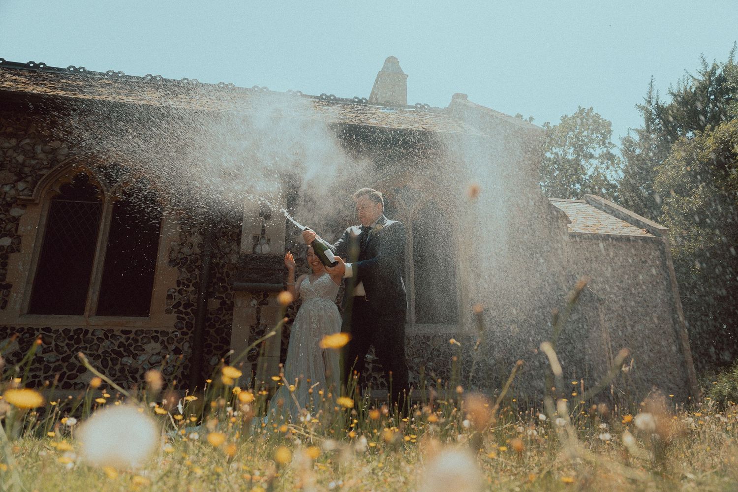 Bride and groom popping champagne in a wildflower meadow outside a rustic stone building on their wedding day.