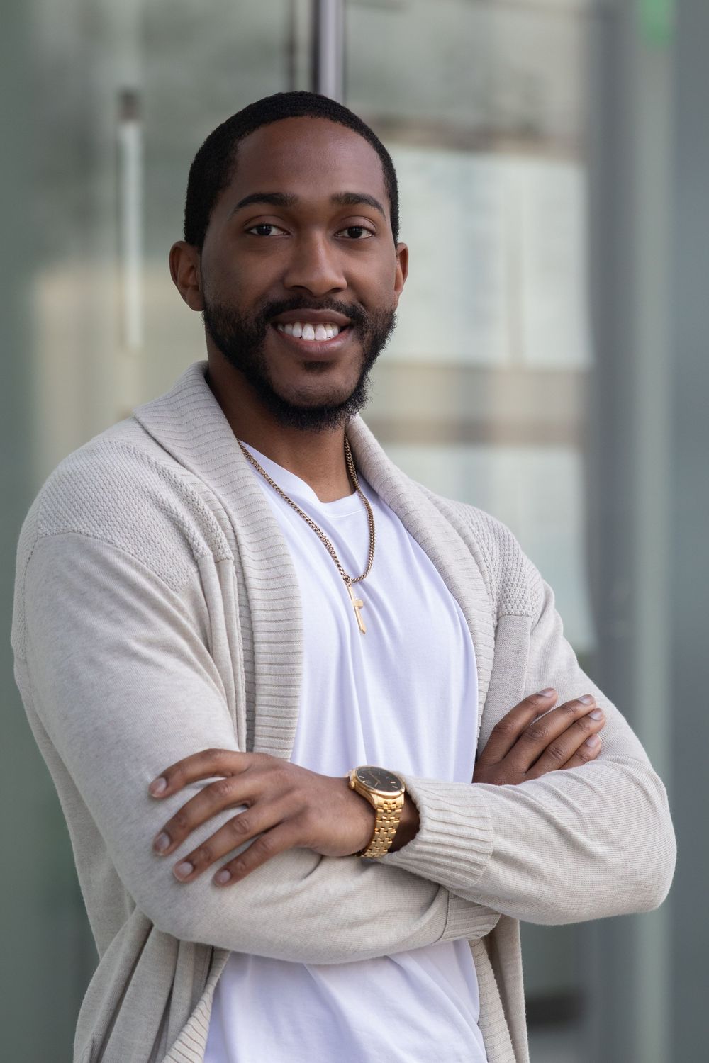 Smiling Black man with beard, arms crossed, wearing white tee, beige cardigan, gold watch and chain necklace outdoors.