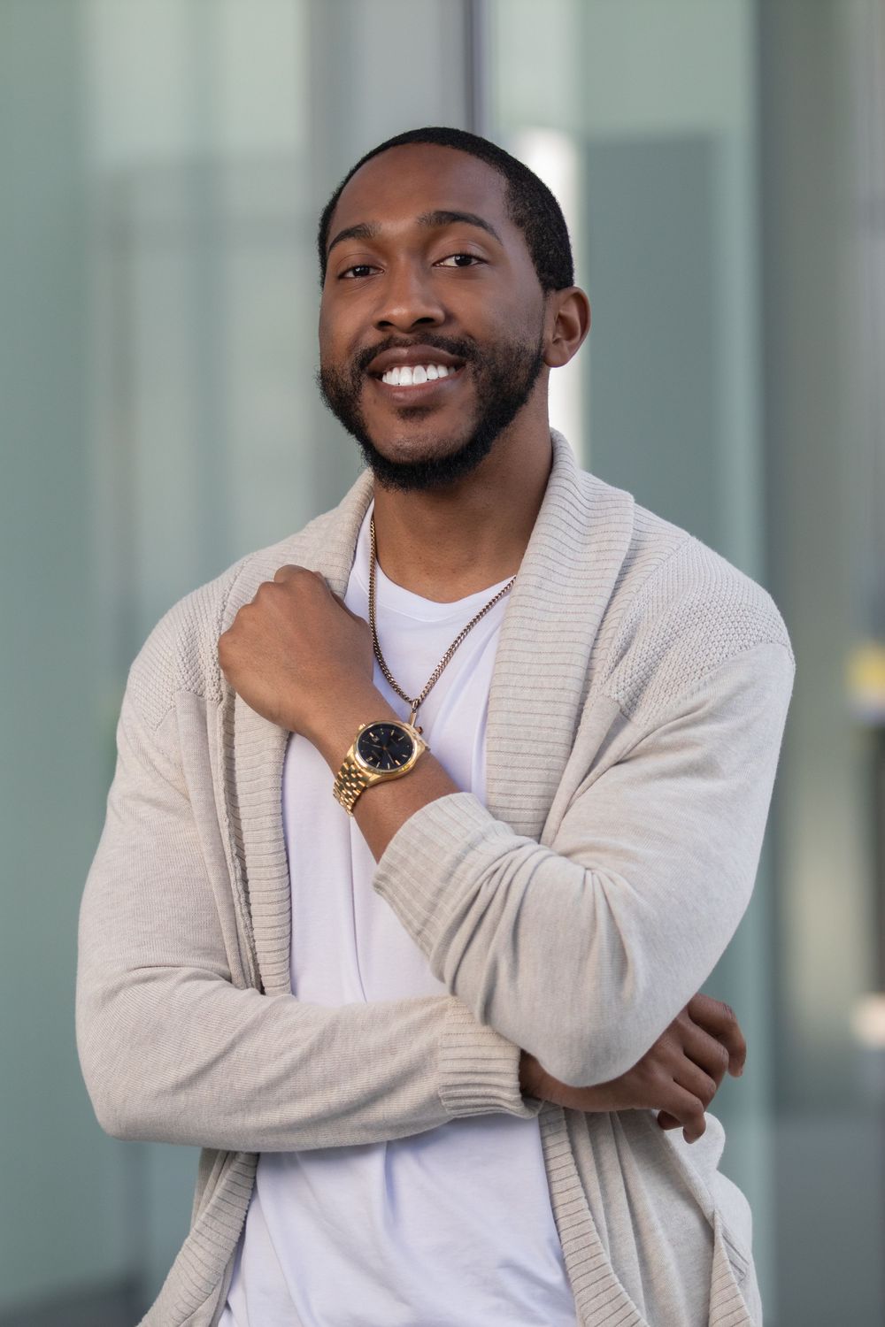 Smiling young Black man in beige cardigan and white tee, wearing gold watch, posing confidently outdoors.