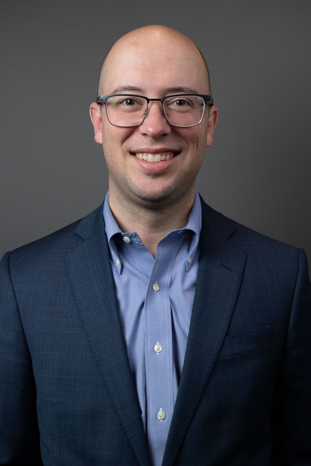 Smiling bald man with glasses wearing a blue dress shirt and navy blazer, professional headshot on gray background.