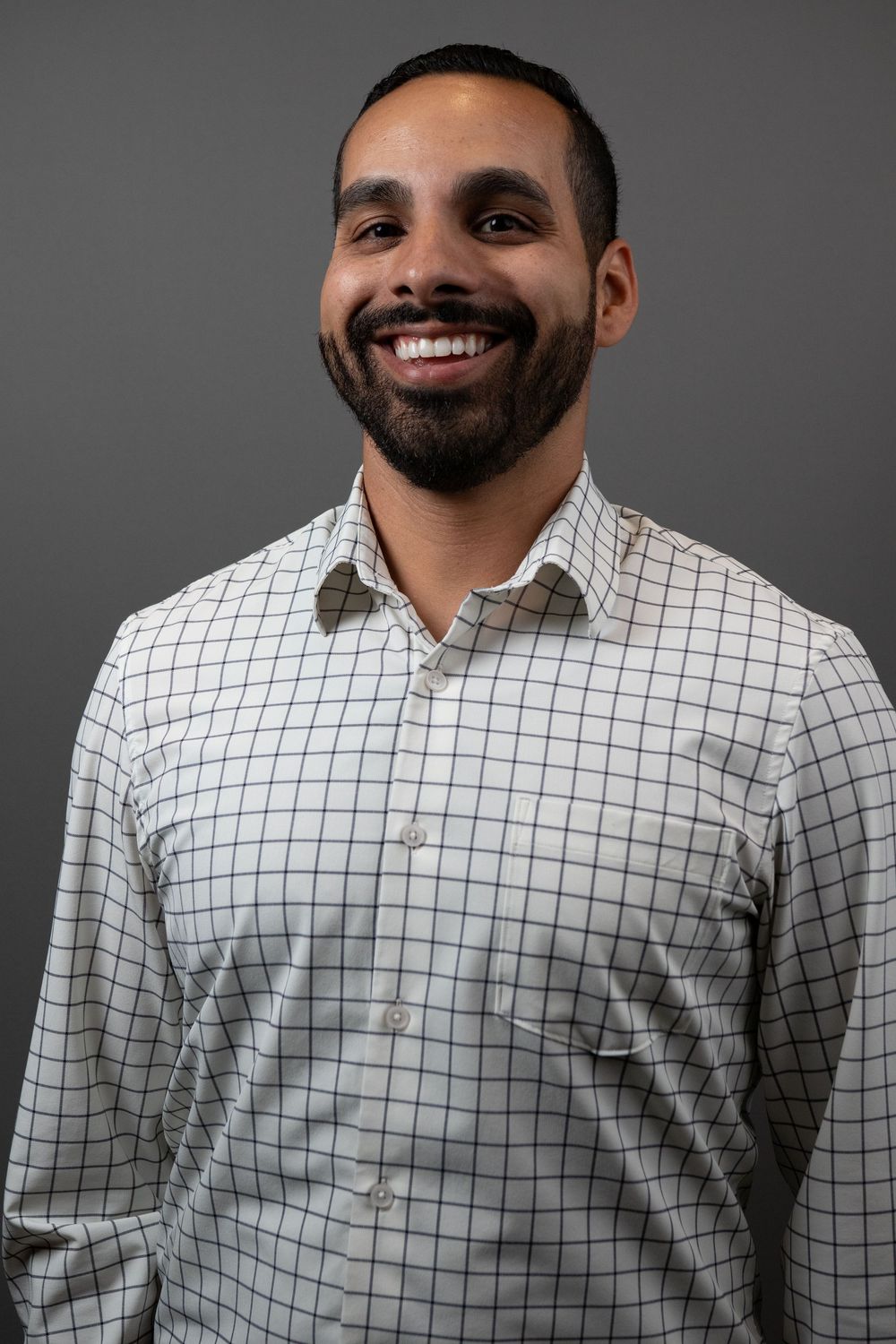 Smiling bearded man in a white checkered button-up shirt posing against a gray background.