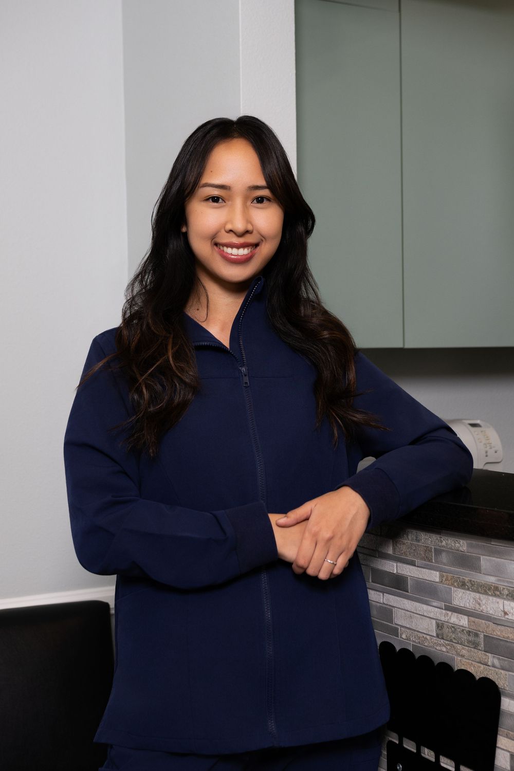 Smiling female healthcare professional in navy blue scrubs jacket posing in a modern medical office.