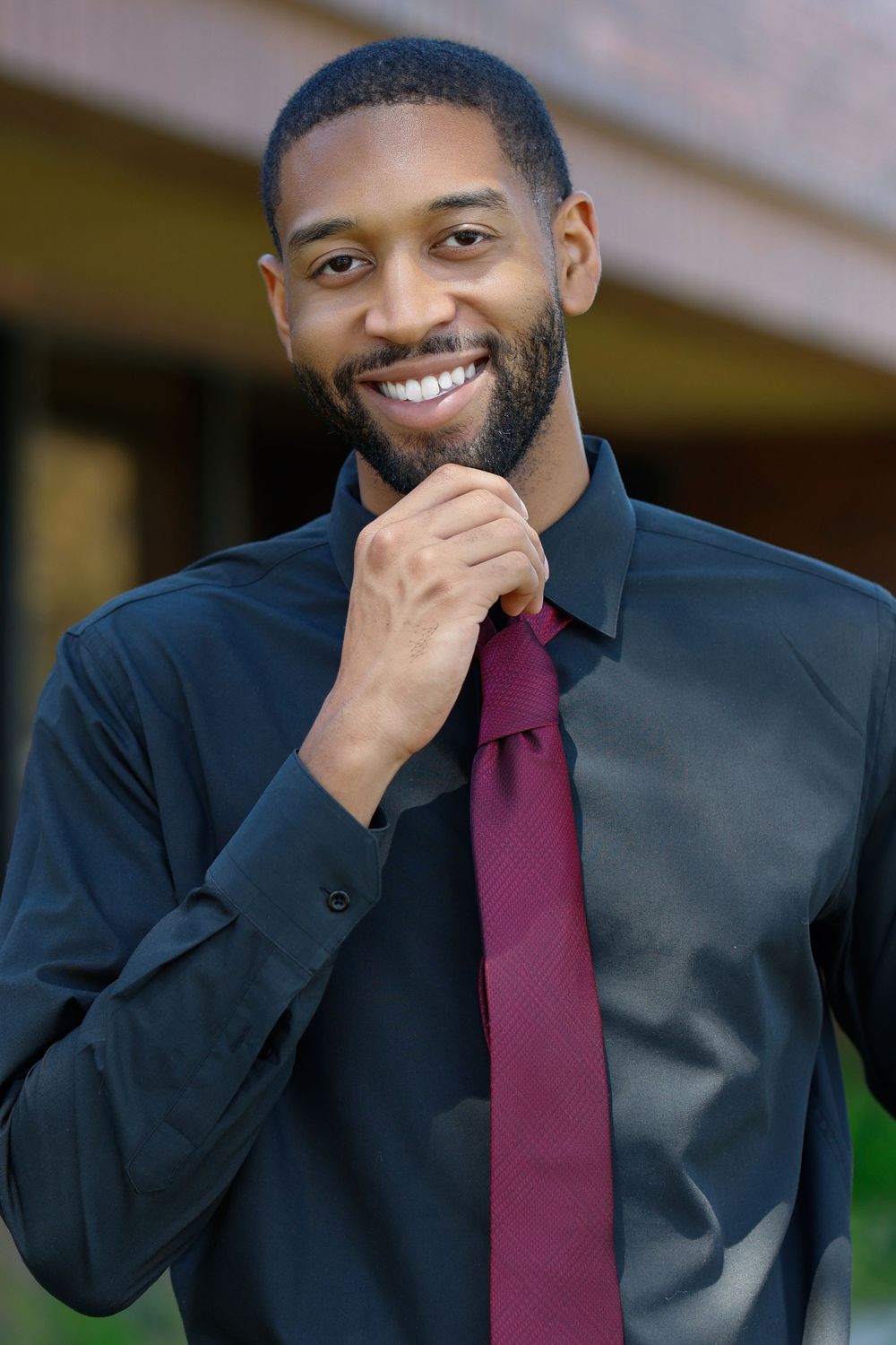 Smiling Black businessman in black shirt and burgundy tie adjusting his collar outdoors.