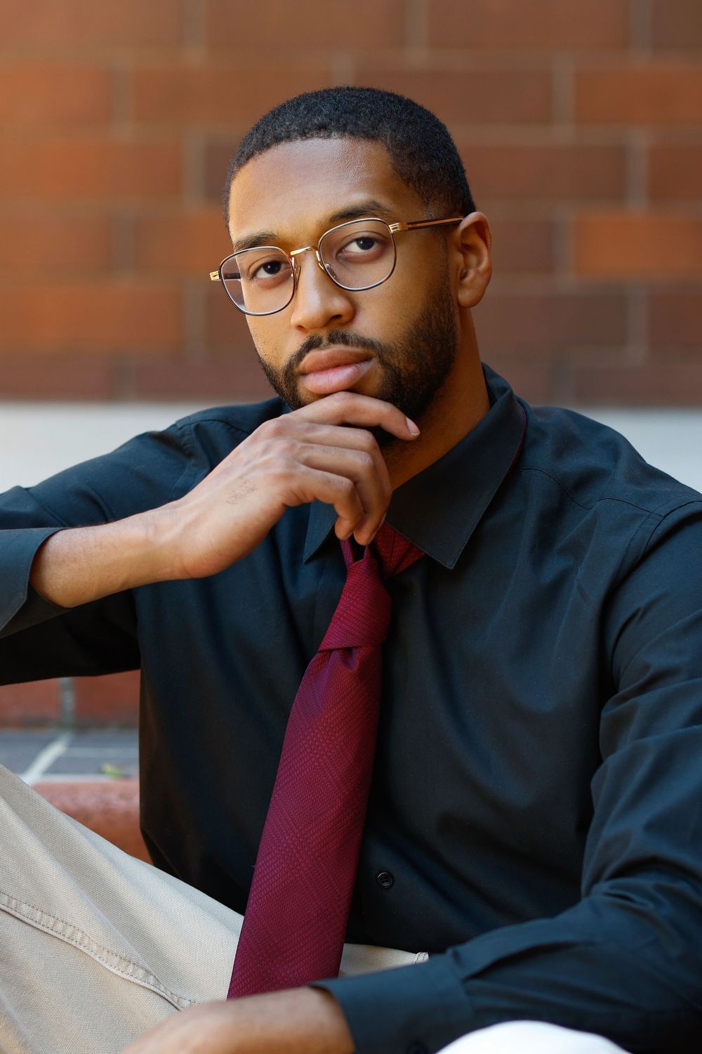 Thoughtful young Black man in black shirt and burgundy tie with glasses, resting chin on hand near brick wall.