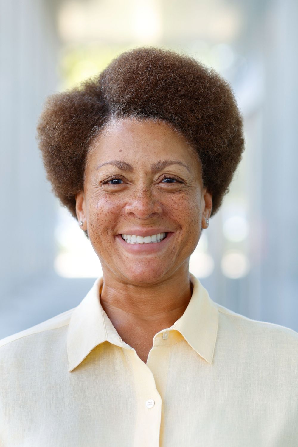 Smiling middle-aged Black woman with natural afro hair wearing a yellow shirt outdoors.