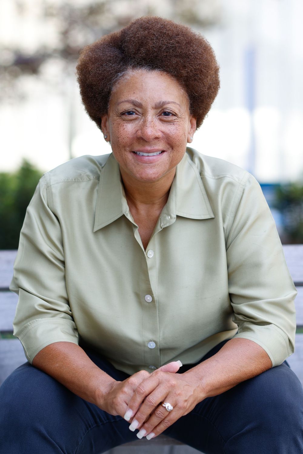 Smiling middle-aged Black woman with afro hair wearing a sage green shirt, sitting outdoors with hands clasped.