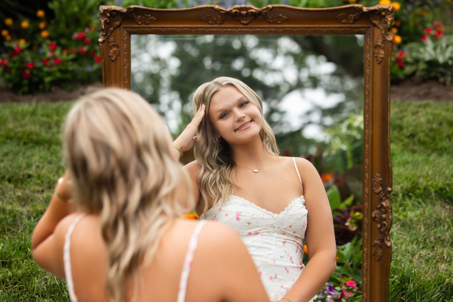 Young blonde woman in floral dress gazes at her reflection in an ornate gold mirror outdoors in a lush garden.