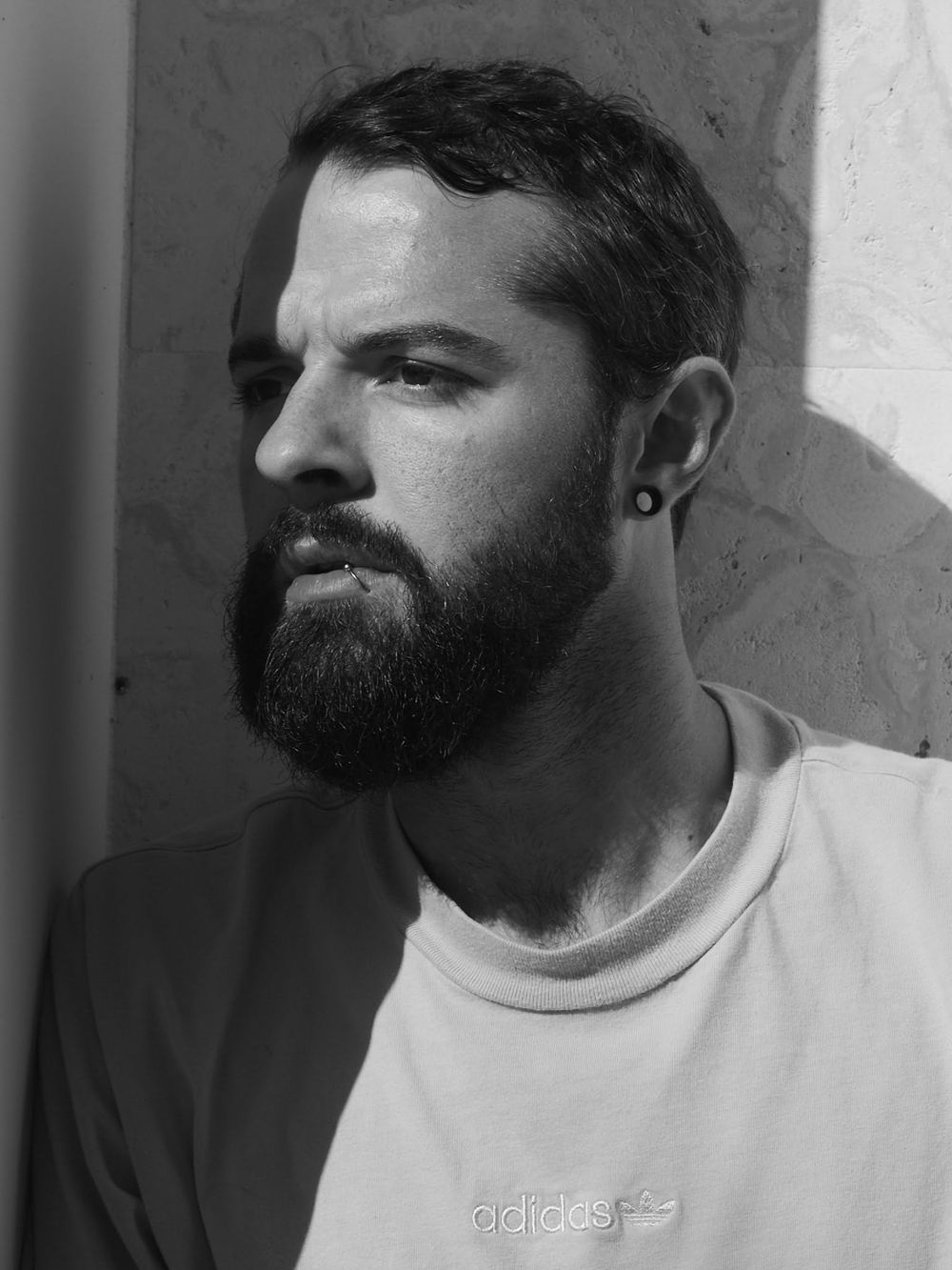 Black and white portrait of a bearded man wearing an Adidas sweatshirt, looking sideways with a small ear stud.