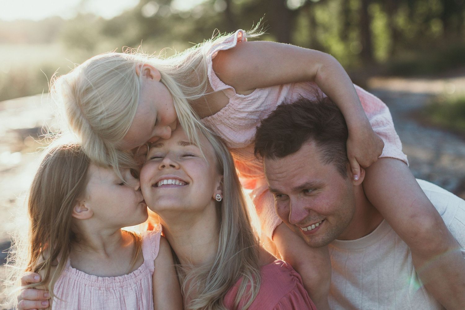 Happy family with two blonde daughters sharing kisses and hugs outdoors in warm sunlight. Mother in focus.