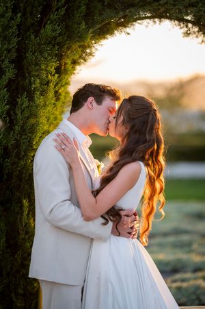 A bride and groom share a romantic kiss at golden hour beneath a lush green archway, glowing in warm sunset light.