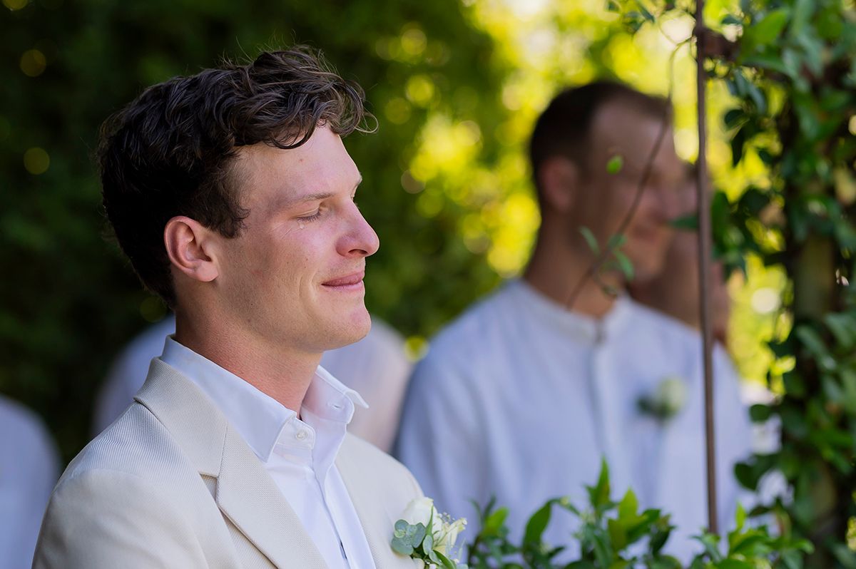 Emotional groom in white suit with boutonniere smiling during outdoor wedding ceremony surrounded by greenery.