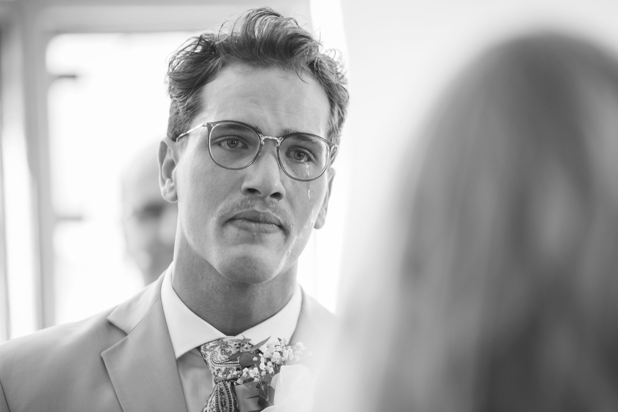 Black and white photo of a groom in glasses and floral boutonniere looking emotional at his wedding ceremony.